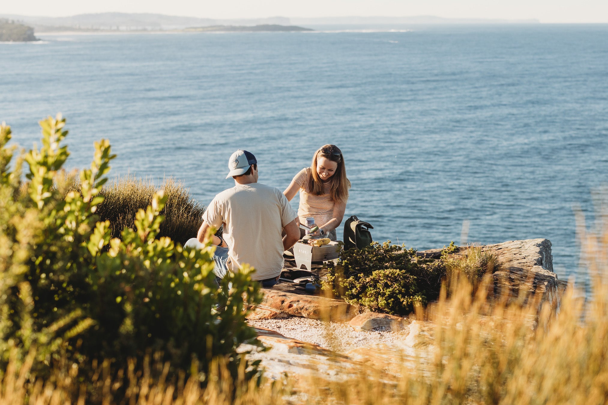 A couple is using portable BBQs for their outdoor cooking in this case the compact STOV BBQ overlooking the ocean.